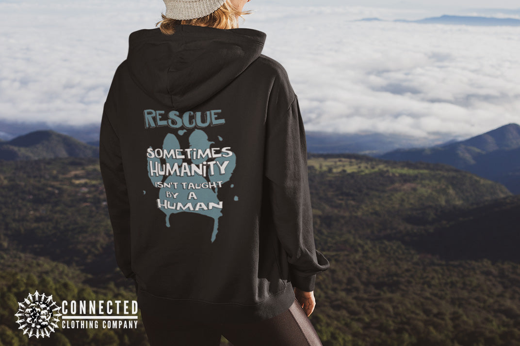 Model wearing Black Show Humanity Unisex Hoodie at the mountains that reads "Rescue. Sometimes humanity isn't taught by a human" - iguanadelabarra - Ethically and Sustainably Made - 10% donated to the Society for the Prevention of Cruelty to Animals